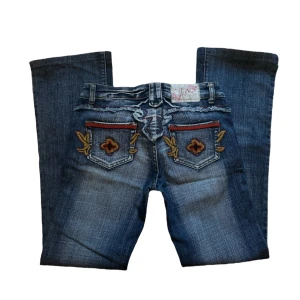 y2k vintage blå jeans med broderade detaljer - Snygga blå jeans med unika broderade detaljer på bakfickorna. Jeansen har en bootcut-stil och är dekorerade med färgglada broderier i rött och gult. Enda är att har lite rip nere vid benslutrt men inte så märkvärdigt. Midjemått: 34 cm rakt över, Innerbelslängd: 78 cm. 💜