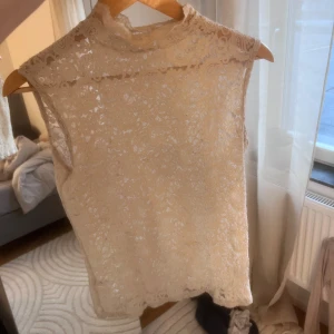 Beige spetsblus - Elegant beige spetsblus med hög krage och ärmlös design. Perfekt för en stilren look med vackert mönster i spetsen 