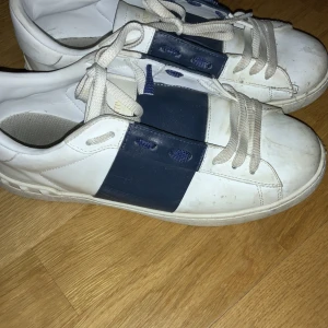 Valentino Garavani sneakers - Snygga Valentino Garavani sneakers i vitt och blått. Skorna har en bred blå rem över ovansidan och klassisk snörning. Perfekta för en stilren look. 