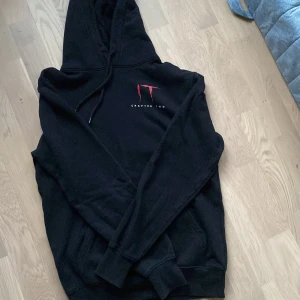 Svart hoodie med IT Chapter Two-tryck - Säljer en svart hoodie med tryck från IT Chapter Two. Den har en stor huva och långa ärmar. På framsidan finns en röd och vit logotyp, och på baksidan ett detaljerat motiv från filmen.
