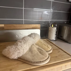 Beige tofflor med fuskpäls - Mysiga beige tofflor med mjuk fuskpäls på insidan och kanten. Perfekta för att hålla fötterna varma under kyliga dagar.