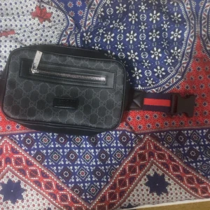 Svart midjeväska från Gucci - Säljer en snygg svart midjeväska från Gucci med klassiskt mönster. Väskan har en dragkedja framtill och ett justerbart bälte i rött och blått. Perfekt för att bära det viktigaste med stil!