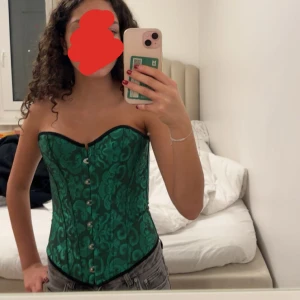Grön korsett med mönster - Snygg grön korsett med ett elegant mönster och knäppning fram. Den har snörning bak för perfekt passform och avtagbara axelband. Perfekt för festliga tillfällen! Passar super bra till halloween, har aldrig använts. 