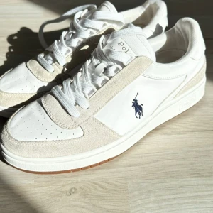 Vita sneakers från Polo Ralph Lauren - Säljer ett par snygga vita sneakers från Polo Ralph Lauren i mycket bra skick. De har en stilren design med mockadetaljer och den klassiska Polo-loggan i blått på sidan. Skorna har snörning och är perfekta för både vardag och fest. Storleken är 40