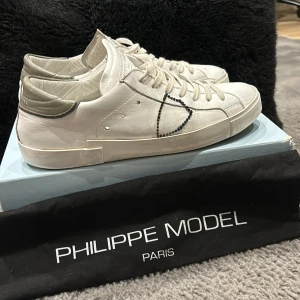 Vita sneakers från Philippe Model - Snygga vita sneakers från Philippe Model med oliv gröna detaljer. Skorna har en stilren design och är mycket trendiga just nu. Box och dustbag tillkommer även. Hör av er vid funderingar!