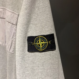 Grå tröja från Stone Island - Säljer en stilren grå tröja från Stone Island med en bröstficka och det ikoniska märket på ärmen. Tröjan har långa ärmar och en avslappnad passform, perfekt för en casual look. Passar inte priser så kom med förslag!