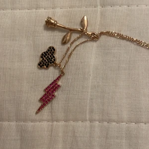 Guldhalsband med berlocker - Unikt guldhalsband med tre berlocker: en ros, en svart musformad berlock och en blixt i rosa stenar. Perfekt för att ge en personlig touch till din stil.