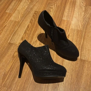 Svarta glittriga stövletter med klack - Snygga svarta stövletter med hög klack och glittrig yta. De har en spetsig tå och dragkedja på sidan för enkel på- och avtagning. Perfekta för att ge en extra touch till din outfit.