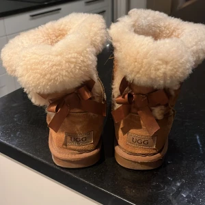 Bruna UGG boots med rosett - Säljer ett par bruna UGG boots med mjukt foder och dekorativa rosetter baktill. Perfekta för kalla dagar med sin varma och bekväma design. Använda max 4 gånger!