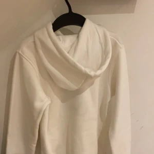 Vit hoodie från Moschino med nallemotiv - Säljer en vit hoodie från Moschino med ett gulligt nallemotiv och text på framsidan. Hoodien har en klassisk design med dragsko i huvan och långa ärmar. Aldrig andvänt har en likadan i svart också!