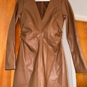 Brun klänning i skinnimitation - Snygg brun klänning i skinnimitation med v-ringning och långa ärmar. Perfekt för höst och vinter med sin stilrena design. Klänningen har en figurnära passform som framhäver silhuetten.