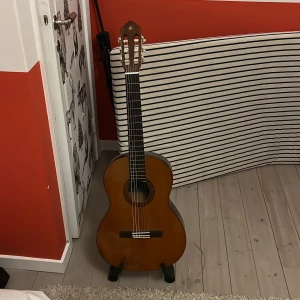 Yamaha CS 40 klassisk gitarr med RockStand ställ - Säljer en Yamaha CS 40 klassisk gitarr, perfekt för nybörjare och unga musiker. Gitarren är tillverkad i Indonesien och har en vacker brun finish med dekorativ rosett runt ljudhålet. Medföljer ett stabilt RockStand gitarrställ. Perfekt för att hålla gitarren säker och lättillgänglig. Passa på att skaffa ett komplett set för att börja spela direkt! 🎸