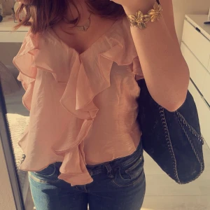 Rosa blus med volangdetaljer - Säljer en superfin rosa blus med volangdetaljer framtill. Perfekt för sommardagar eller en festlig kväll. Blusen har en luftig passform och är tillverkad i ett lätt och bekvämt material. Passar perfekt med jeans eller kjol för en chic look!