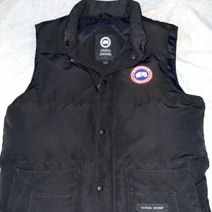 Svart dunväst från Canada Goose - Säljer en svart dunväst från Canada Goose i mycket bra skick. Västen har en klassisk design med knappar och en hög krage för extra värme. Perfekt för höst och vinter, och den passar bra till både vardag och utomhusaktiviteter. Storleken är herr M.