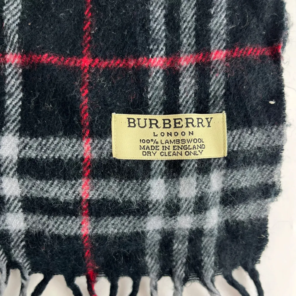 Säljer en klassisk rutig halsduk från Burberry i 100% lammull. Den har en snygg kombination av svart, grått och rött med fransar i ändarna. Perfekt för att hålla sig varm och stilren under kyliga dagar. Passar både höst och vinter!. Övrigt.