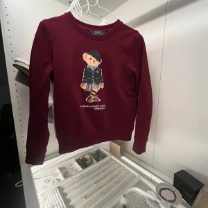 Vinröd tröja från Ralph Lauren - Säljer en superfin vinröd tröja från Ralph Lauren med en söt björn på framsidan. Tröjan är långärmad och perfekt för höst och vinter. Den är i mycket bra skick och passar perfekt för en casual look. Perfekt för dig som älskar stilrena och bekväma kläder! 🍂🐻