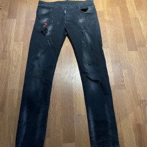Svarta jeans från Dsquared2 - Säljer ett par svarta jeans från Dsquared2 i bra skick. De har en cool distressed look med slitningar och en röd logga på baksidan. Perfekta för en avslappnad men stilren outfit. Midwaist passform och dragkedja framtill. Äkta!