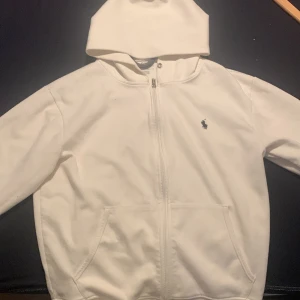 Vit huvtröja från Ralph Lauren - Säljer en snygg vit huvtröja från Ralph Lauren i mycket bra skick. Den har en dragkedja framtill och en liten broderad logga på bröstet. Perfekt för en casual look eller kyliga dagar. Storleken är normal och materialet känns mjukt och bekvämt.