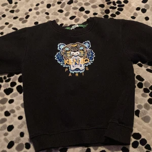 Svart sweatshirt från Kenzo - Säljer en svart sweatshirt från Kenzo med ett coolt tigerbroderi på framsidan. Tröjan har långa ärmar och är i mycket bra skick. Perfekt för höst och vår när du vill ha något stilrent men ändå iögonfallande. Passar både till vardags och festliga tillfällen!