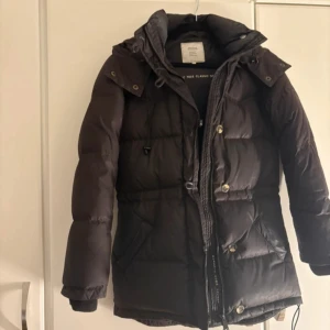 dunjacka från Boomerang - Säljer en dunjacka från Boomerang med huva och knappar. Jackan har en klassisk design med quiltade detaljer och en liten logotyp på ärmen. Perfekt för kyliga dagar. Använd men i bra skick!