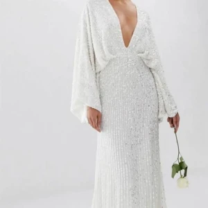 Vit glittrig balklänning med släp  - Säljer min fina balklänning som är slutsåld på hemsidan. Det är en vit långklänning med glittrande detaljer och djup v-ringning. Den är endast använd en gång och sitter som en M. Klänningen har långa, vida armar och ett fint släp där bak🥰