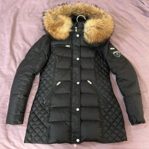 Svart Rockandblue dunjacka - Säljer en snygg svart dunjacka från Rockandblue i nyskick. Köpt för 4000kr, använd 2 gånger! Jackan har en avtagbar luva med äkta päls och dragkedja framtill. Perfekt för kalla vinterdagar! 