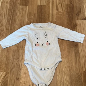 Vit långärmad babybody från Next - Söt vit babybody från Next med ett kaninansikte tryckt på framsidan. Bodyn har långa ärmar och knäppning i grenen för enkel påklädning. Perfekt för de små under vår och sommar.