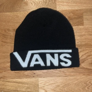 Svart mössa från Vans - Säljer en svart stickad mössa från Vans i mycket bra skick. Den har en bred uppvikt kant med det ikoniska Vans-loggan i vitt. Perfekt för att hålla sig varm och stilren under kalla dagar. Passar både till vardags och för skateparken!