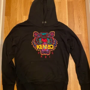 Svart hoodie från Kenzo - Säljer en svart hoodie från Kenzo med ett färgglatt broderat tigerhuvud och texten 'Kenzo Paris' på framsidan. Den har en klassisk passform med långa ärmar och en praktisk magficka. Perfekt för både vardag och chill, saknar huvans rep för att ändra storleken . Skriv till mig innan ni köper. 
