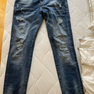 Blå jeans från Dsquared2 - Säljer ett par snygga blå jeans endast provade  från Dsquared2 i storlek 33. De har en cool slitningseffekt och en klassisk femficksdesign. Perfekta för en avslappnad stil med en touch av edge. Passar både till vardags och fest!