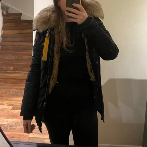 Svart parajumpers med pälsdetaljer - Säljer en snygg svart parkajacka med stor päls på huvan. Jackan har långa ärmar och en gul detalj vid dragkedjan. Perfekt för kalla vinterdagar och håller dig varm och stilren. Passar bra till både vardag och fest.