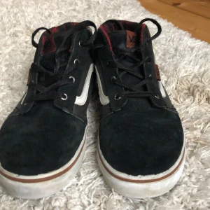Svarta sneakers från Vans - Säljer ett par svarta Vans sneakers i bra skick. De har en klassisk vit sula och den ikoniska vita sidoremsan. Insidan är fodrad med röd och svart rutig textil, vilket ger en cool kontrast. Perfekta för vardagsbruk och streetwear-stil!