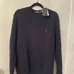 Snygg mörkblå stickad tröja från Polo Ralph Lauren med kabelstickat mönster. Tröjan har en klassisk rund hals och det ikoniska röda logotypbroderiet på bröstet. Perfekt för en stilren look.