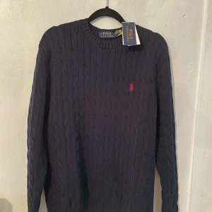 Mörkblå stickad tröja från Polo Ralph Lauren - Snygg mörkblå stickad tröja från Polo Ralph Lauren med kabelstickat mönster. Tröjan har en klassisk rund hals och det ikoniska röda logotypbroderiet på bröstet. Perfekt för en stilren look.