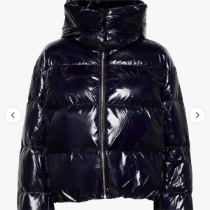 Svart pufferjacka från Tommy Hilfiger - Snygg svart pufferjacka från Tommy Hilfiger med glansig yta och dragkedja framtill. Jackan har en hög krage och en stor huva för extra värme. Perfekt för kyliga dagar.