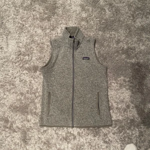 Grå fleeceväst från Patagonia - Säljer en stilren grå fleeceväst från Patagonia med dragkedja framtill. Västen har en normal passform och är perfekt för lager-på-lager under kyligare dagar. Den har två praktiska fickor med dragkedja.