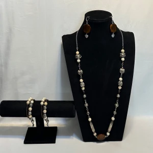 Set med halsband, armband och örhängen - Snyggt smyckeset med ett långt halsband, matchande armband och örhängen. Halsbandet har pärlor i olika storlekar och färger, inklusive silver och brunt. Armbanden är elastiska med liknande pärlor. Örhängena har stora, runda bruna hängen. Perfekt för att ge en bohemisk touch till din outfit! Ett par örhängen och ett par armband plus en halsband handgjorda mycket mycket fint nytt oanvänd 