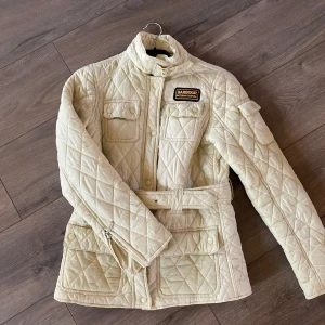 Beige quiltad jacka från Barbour - Säljer en snygg beige quiltad jacka från Barbour i mycket bra skick. Jackan har ett bälte i midjan och flera praktiska fickor med knappar. Den är långärmad och perfekt för höst och vår. Klassisk och stilren design som passar till det mesta!