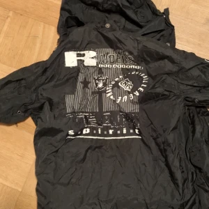 Äkta vintage Svart NFL Raiders jacka från Campri, tidigt 90-tal - Säljer äkta 90's NFL Raiders jacka från Campri med tryck på ryggen och ärmen. Jackan har en huva och knappar framtill. Perfekt för fans av NFL och Raiders. Officiellt licensierad produkt.