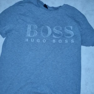 Grå t-shirt från Hugo Boss - Snygg grå t-shirt från Hugo Boss med stort BOSS-tryck på framsidan. Perfekt för en stilren och avslappnad look. Kortärmad och tillverkad i mjukt material för extra komfort.