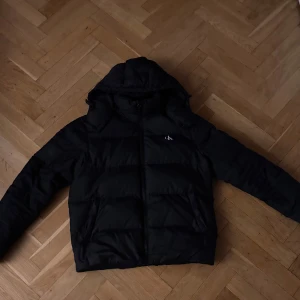 Svart pufferjacka från Calvin Klein Jeans - Snygg svart pufferjacka från Calvin Klein Jeans med huva och dragkedja. Perfekt för kyliga dagar med sin vadderade design. Diskret CK-logga på bröstet och ärmen ger en stilren touch.