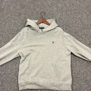 Grå hoodie från Ralph Lauren - Säljer en stilren och bekväm grå hoodie från Ralph Lauren. Den har en klassisk passform med långa ärmar och en praktisk magficka. Perfekt för både vardag och avslappnade tillfällen. Den lilla loggan på bröstet ger en extra touch av stil. Passar perfekt till jeans eller joggers!