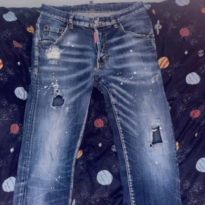 D2QUARED2 jeans - Säljer ett par coola jeans med slitningar och färgstänk. De har en normal passform och är perfekta för en avslappnad stil. Jeansen har en klassisk blå färg och är i bra skick trots slitningarna. Perfekta för höst och vår!