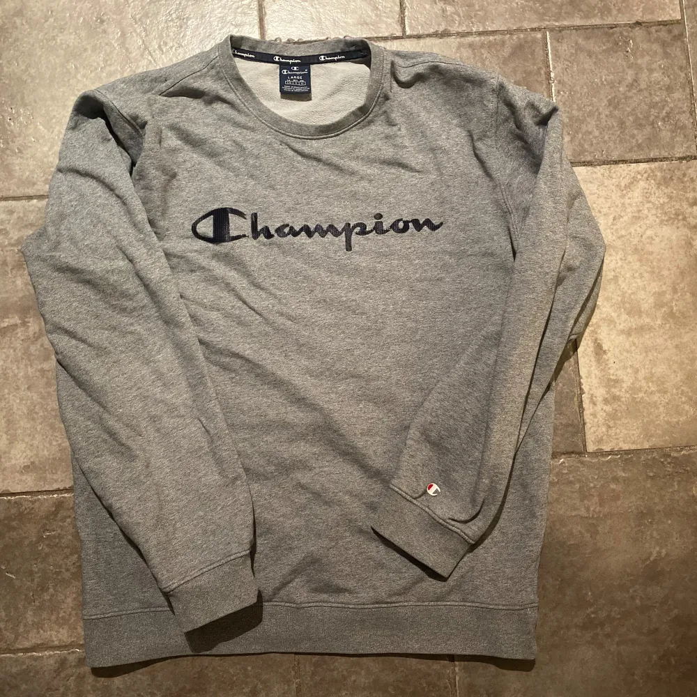 Säljer en grå sweatshirt från Champion i bra skick. Den har ett stort broderat Champion-logga på bröstet som delvis har börjat lossna lite samt en liten logga på ärmen. Perfekt för höst och vår, med långa ärmar och en bekväm passform. Skön och stilren för vardagsbruk! . Tröjor & Koftor.