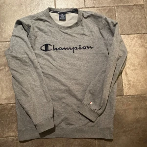 Grå sweatshirt Champion - Säljer en grå sweatshirt från Champion i bra skick. Den har ett stort broderat Champion-logga på bröstet som delvis har börjat lossna lite samt en liten logga på ärmen. Perfekt för höst och vår, med långa ärmar och en bekväm passform. Skön och stilren för vardagsbruk! 