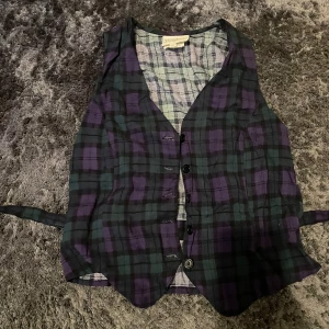 Rutig väst med knappar - Säljer en cool rutig väst i lila och grön med knappar framtill. Perfekt för att ge din outfit en extra edge. Västen har ett klassiskt snitt och är i bra skick. Passar både till vardags och fest!