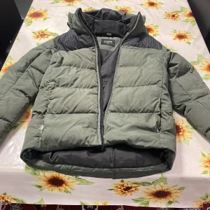 Grön pufferjacka från Everest - Säljer en grön pufferjacka från Everest i bra skick och passar S-M. Jackan har en dragkedja framtill och är perfekt för kalla dagar. Den är quiltad och har en huva för extra värme. Passar perfekt för höst och vinter!