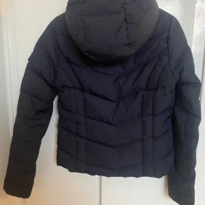 Marinblå pufferjacka från Hollister - Säljer en svart pufferjacka från Hollister i bra skick. Jackan har en dragkedja framtill och en mysig huva. Perfekt för kyliga dagar och har en stilren design med Hollisters logga på bröstet. Passar både till vardags och lite finare tillfällen.