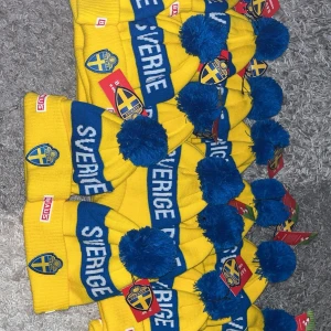 Gul och blå mössa med Sverige-märke - Snygg gul och blå mössa med texten 'Sverige' och ett stort blått tofs på toppen. Perfekt för att visa ditt stöd för Sverige. Märket 'AUS' syns på kanten.