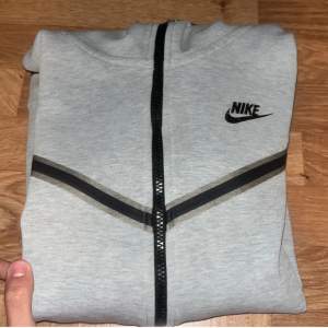 Säljer en grå hoodie från Nike i bra skick. Den har en dragkedja framtill och en snygg svart detalj över bröstet. Perfekt för höst och vår, och passar både till träning och vardag. Storlek S, men passar även om du gillar en lite tightare passform.